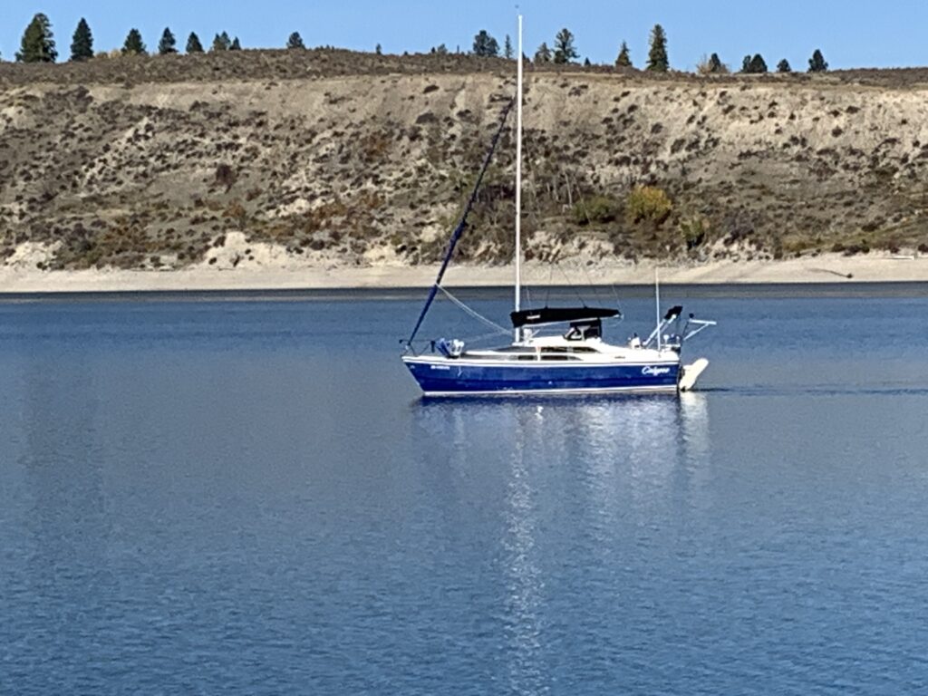 Image of a sailboat cruising on a blue water lake with a hill and trees in the background.